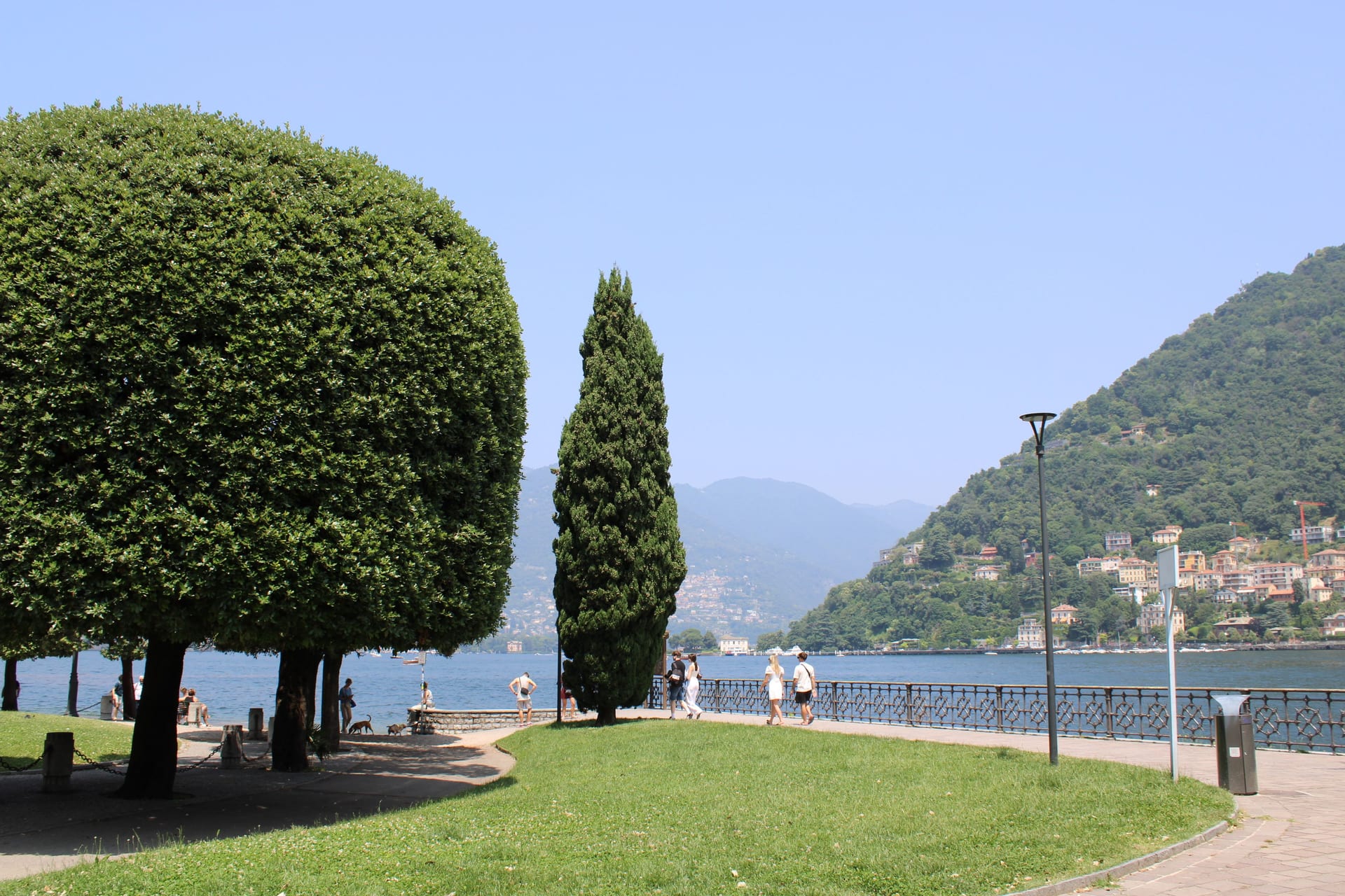 Une pelouse arborée et une promenade au bord du lac à Côme, avec un village coloré sur l'autre rive