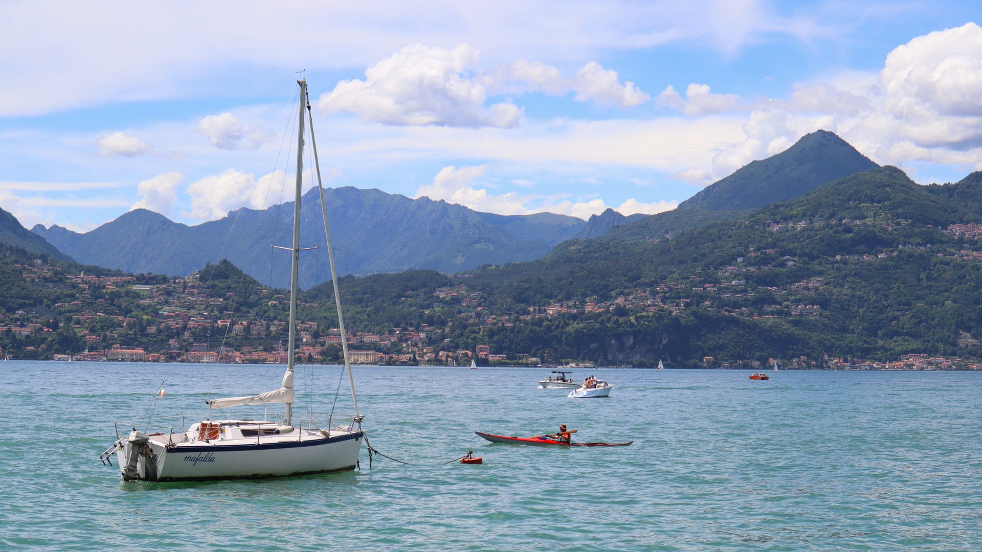 Ein Segelboot und ein Kajak auf dem Comer See an einem Sommertag, im Hintergrund ein Dorf am Ufer