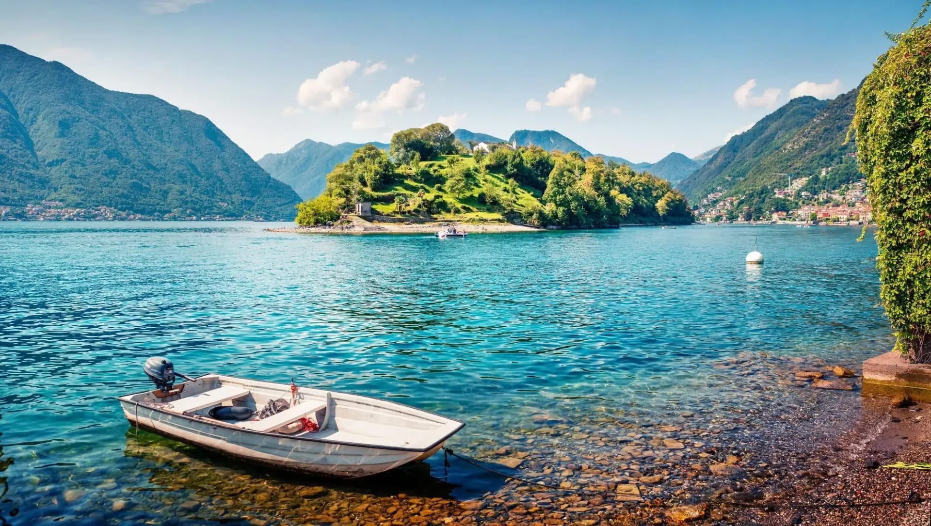 L'Isola Comacina vista dalla riva della Tremezzina: una barca bianca ormeggiata nell'acqua trasparente, l'isola verde al centro del lago e le montagne sullo sfondo