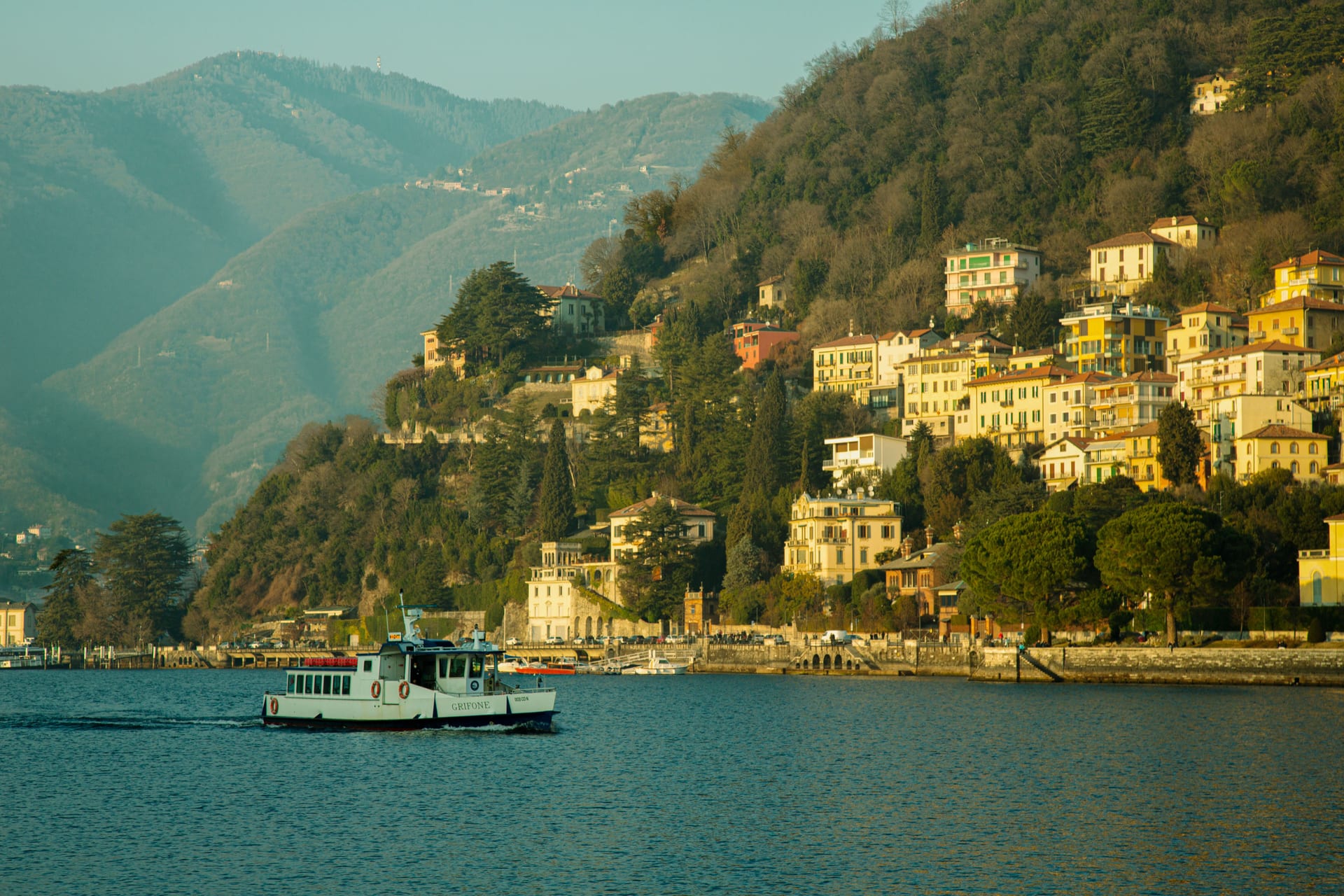 Ein kleines Passagierboot fährt am Comer See vor einem Dorf mit bunten Häusern am Hang vorbei