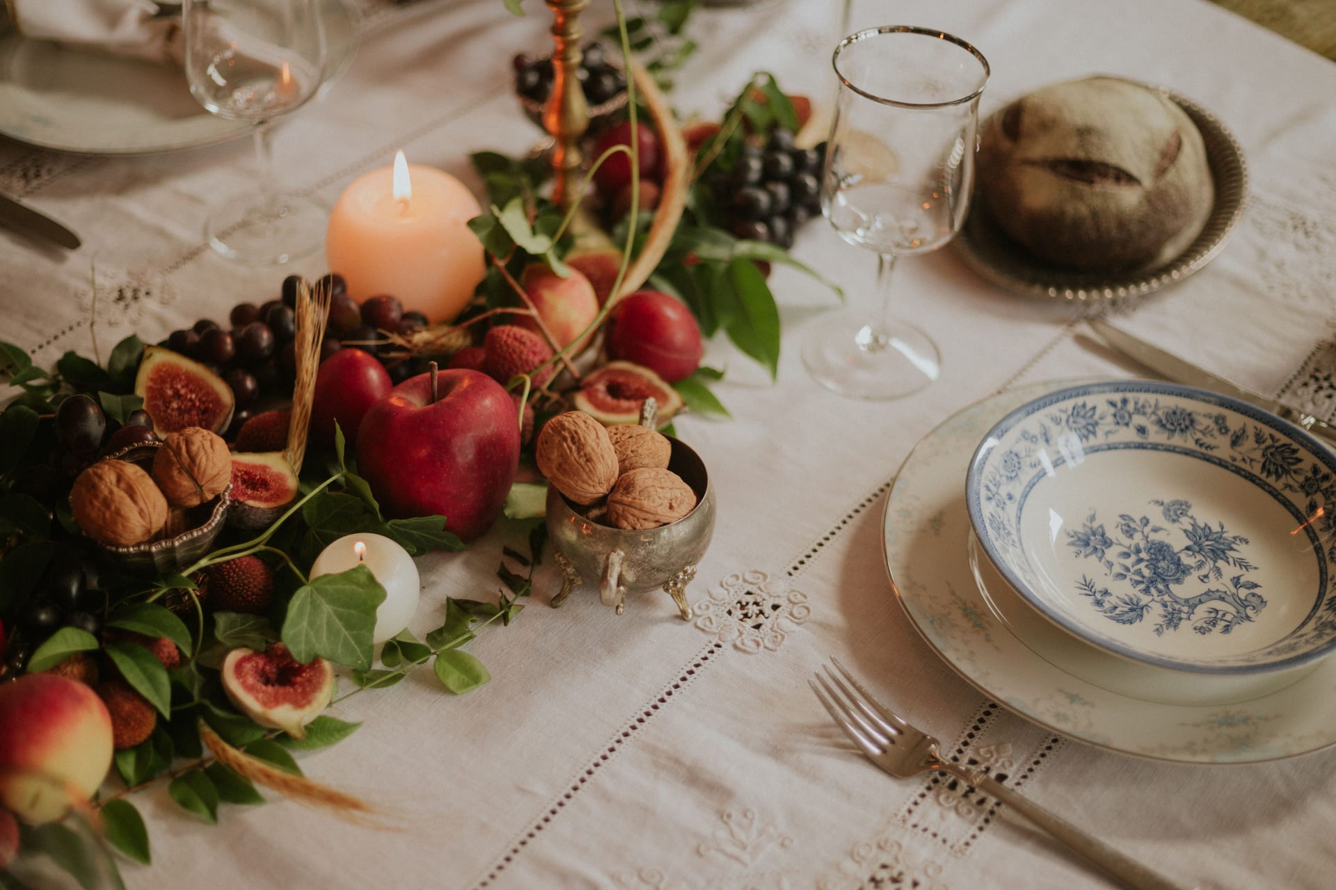 Gedeckter Tisch mit bestickter Leinendecke, gemustertem Porzellan, brennenden Kerzen und Herbstfrüchten: Feigen, Äpfel, Trauben und Walnüsse