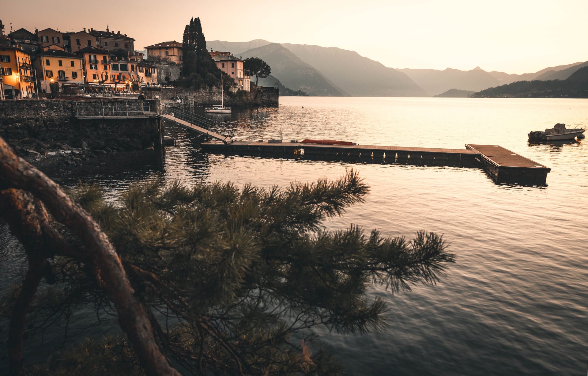 Un molo di legno che si allunga nel lago al tramonto, con un ramo di pino in primo piano e le case color pastello di un borgo sulla riva opposta