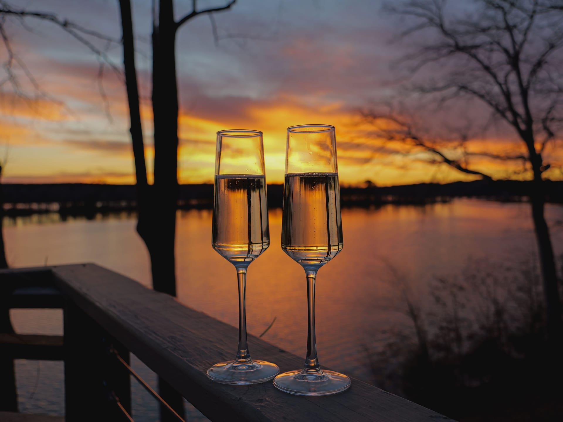 Two flutes of sparkling wine on a wooden railing, in front of a lake at sunset