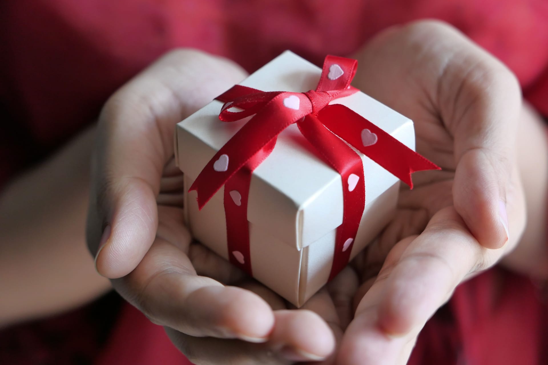 Two hands holding a small white gift box tied with a red ribbon printed with hearts